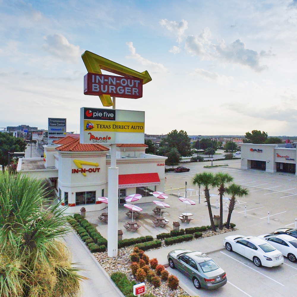 Aerial view of an In-N-Out Burger restaurant with outdoor seating, palm trees, parked cars, and nearby businesses on a partly cloudy day.